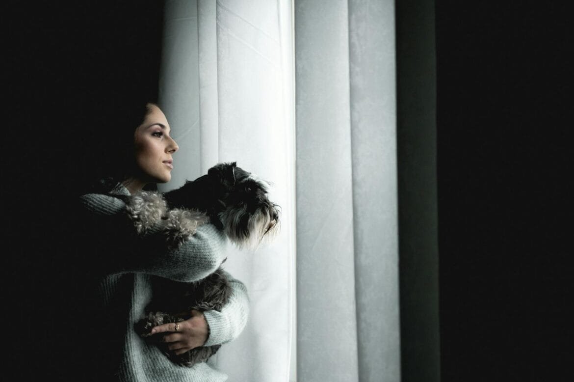 A woman in a sweater holds a dog by a window, gazing outside in soft lighting.