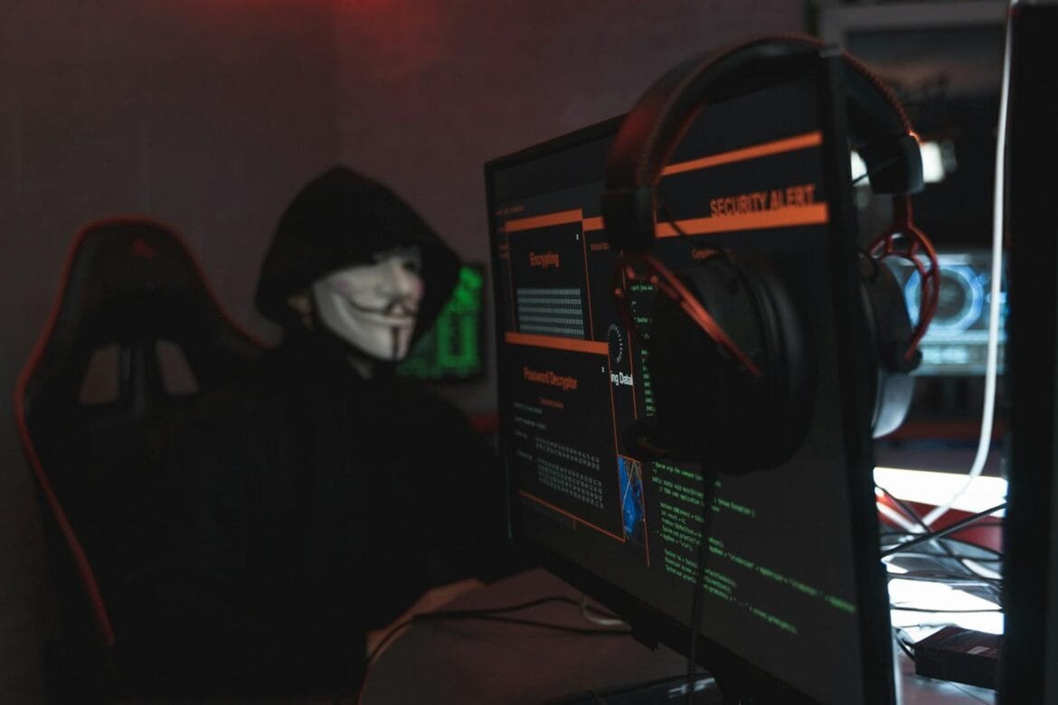 A person wearing a Guy Fawkes mask engaged in hacking activities on a computer in a dimly lit room.