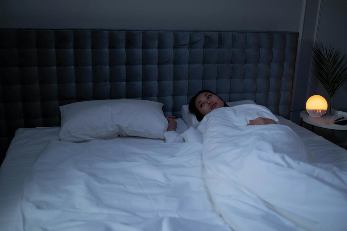 An Asian woman lying awake under soft white bedding in a dimly lit modern bedroom.