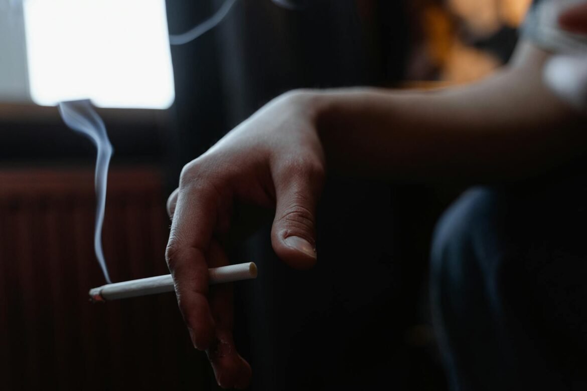 Close-up of a hand holding a lit cigarette indoors with smoke rising in a dimly lit room.