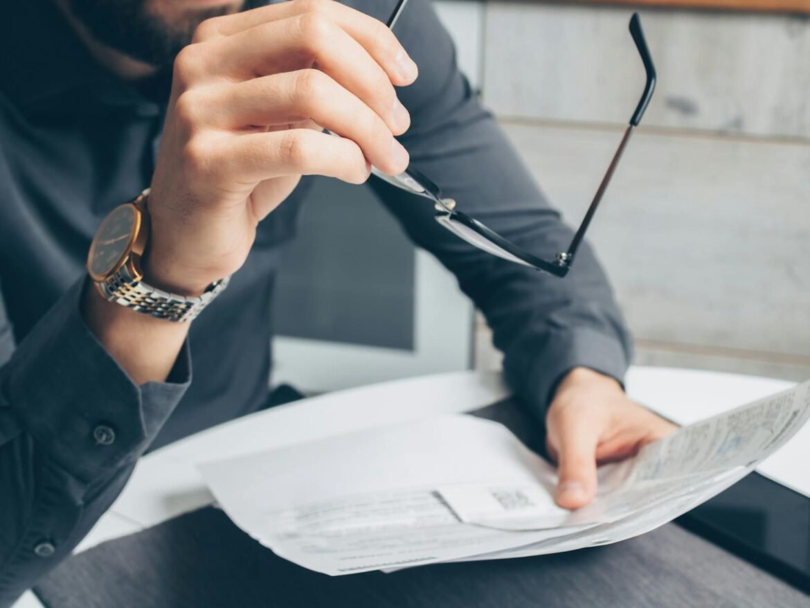 Photo by Mikhail Nilov Man in formal attire reviewing paperwork, holding glasses. Business setting.