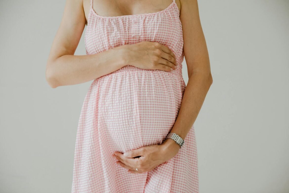 Photo by RDNE Stock project Close-up of pregnant woman holding her belly, wearing a pink checkered dress indoors.