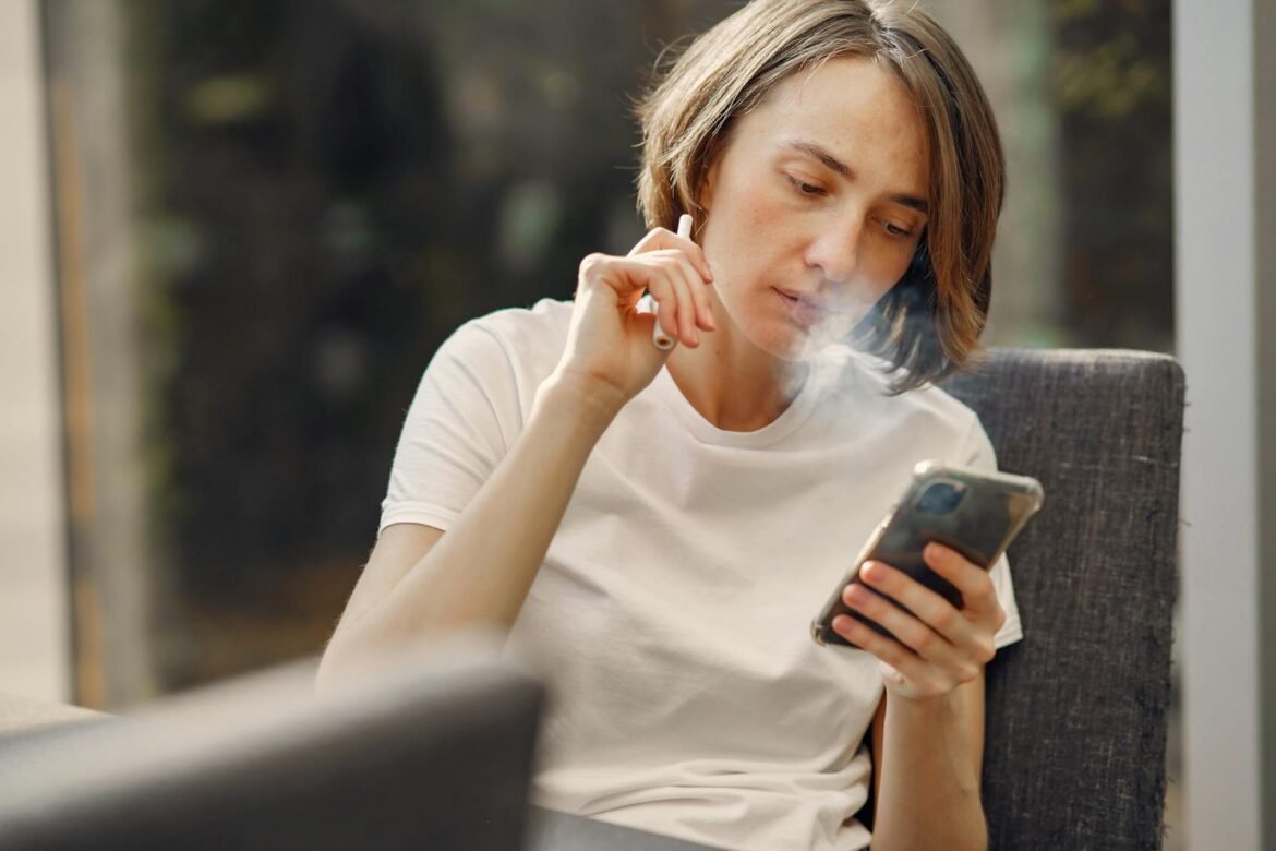 A woman sits indoors, using a smartphone while vaping, creating a relaxed atmosphere.