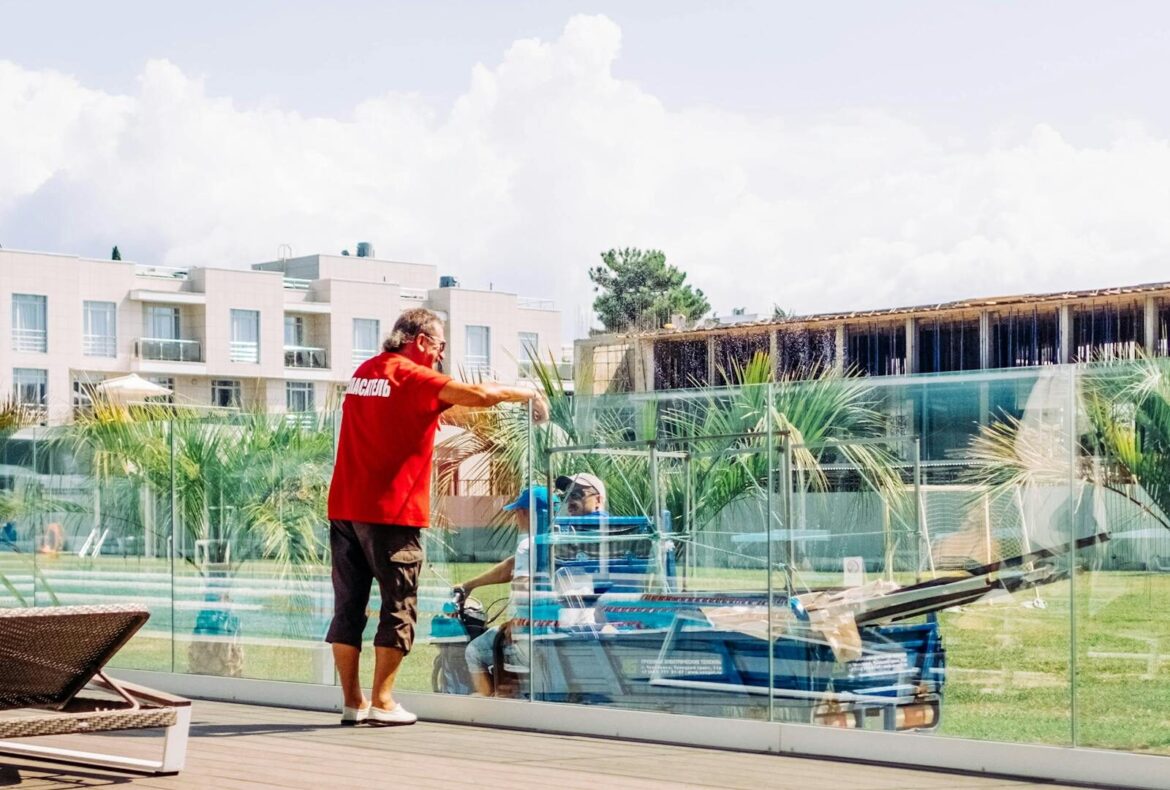 A man fixing glass railing near modern buildings in a sunny outdoor area.